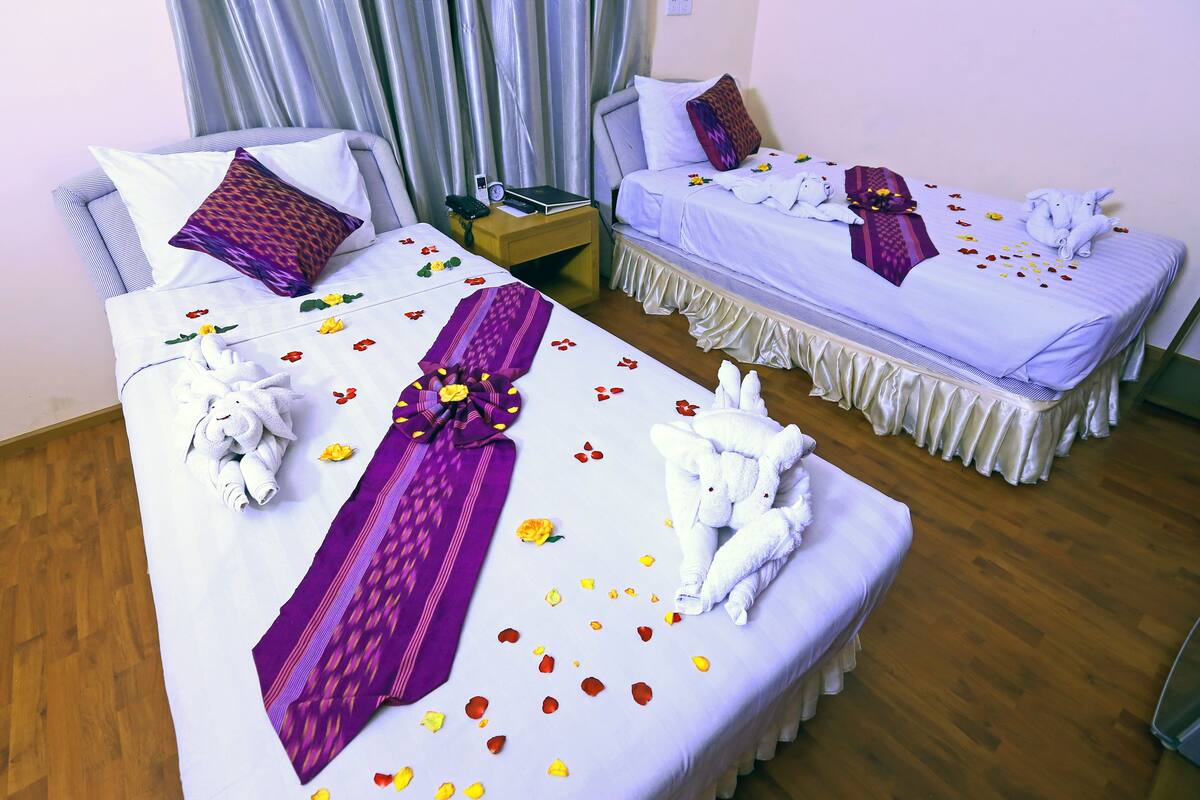 Dos camas ordenadas con toallas y almohadones decorativos, una mesita de noche con un libro y un control remoto, y una cortina al fondo.