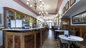 Een bar met houten toonbanken, een kroonluchter, ingekaderde foto’s aan de muur en een zithoek met tafels en stoelen.