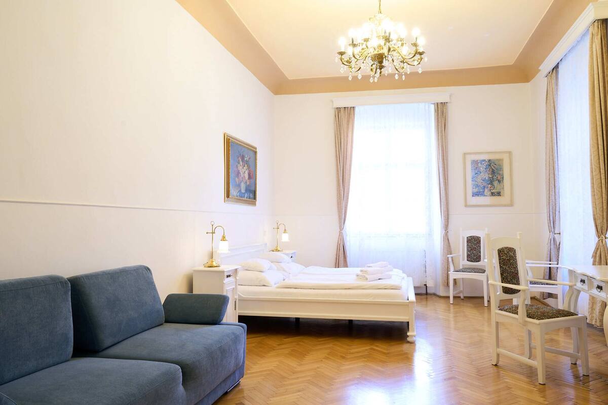 A hotel room with a bed, a blue sofa, a white chair, and a chandelier.