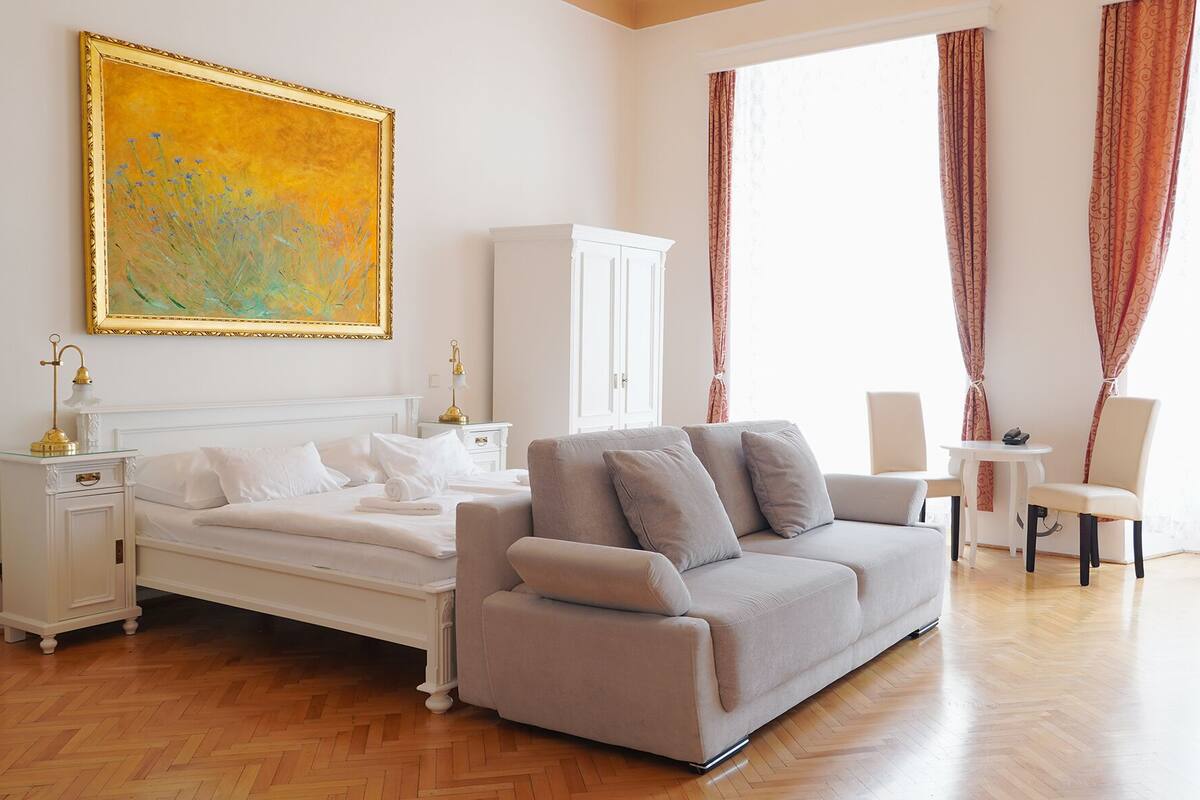 A hotel room with a bed, a sofa, a white cabinet, a painting, and a window with curtains.