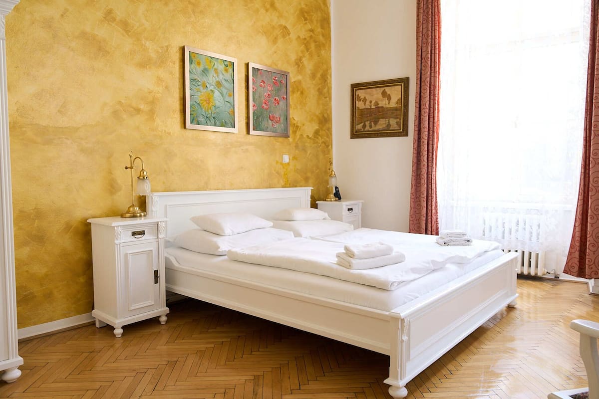A hotel room with a large bed, white nightstands, a bedside table with a lamp, and artwork on the walls.