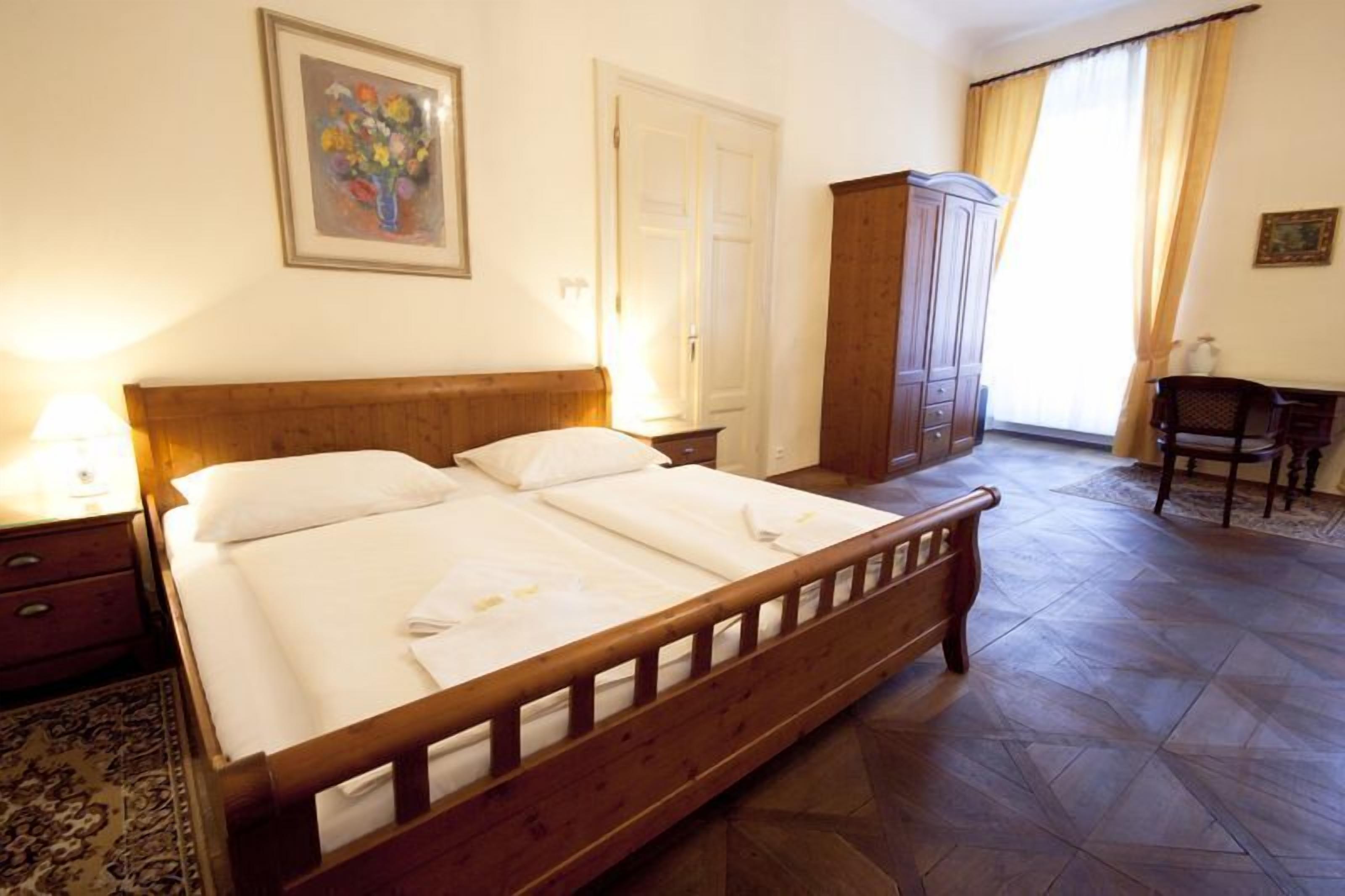 Ein ordentlich bezogenes Bett mit Holz-Kopfteil, ein Nachttisch mit Lampe, ein Kleiderschrank und ein Stuhl in einem gut beleuchteten Zimmer mit Fliesenboden.