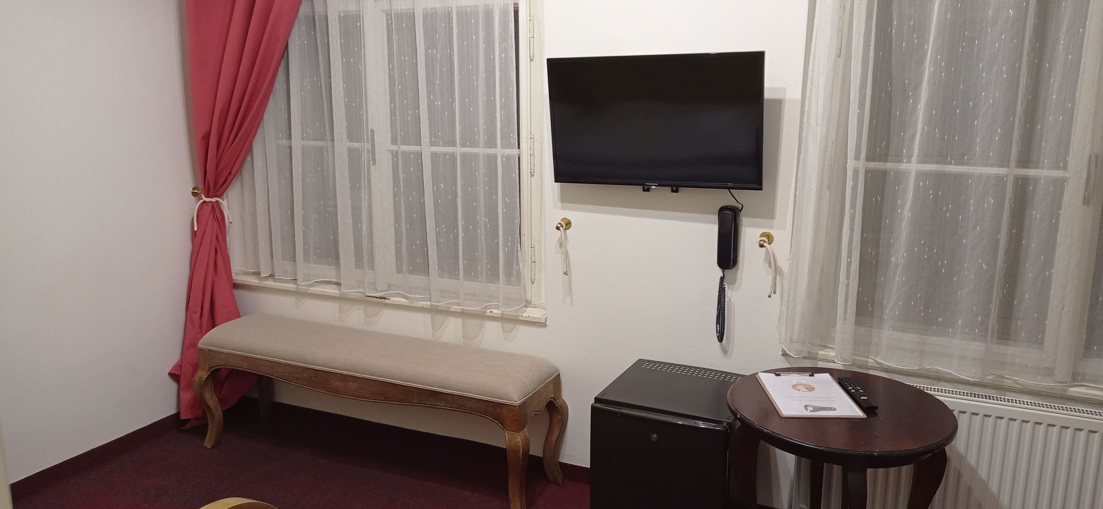 Una habitación con un banco, una pequeña mesa con un documento, un altavoz negro y un televisor montado en la pared.