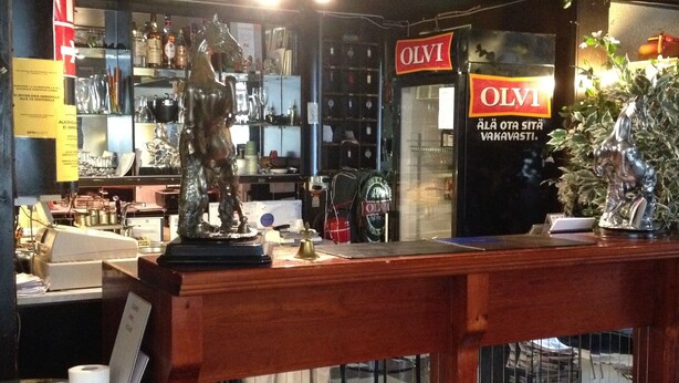 Uma área de bar com balcão de madeira, prateleiras com garrafas e um letreiro com a inscrição "OLVI".