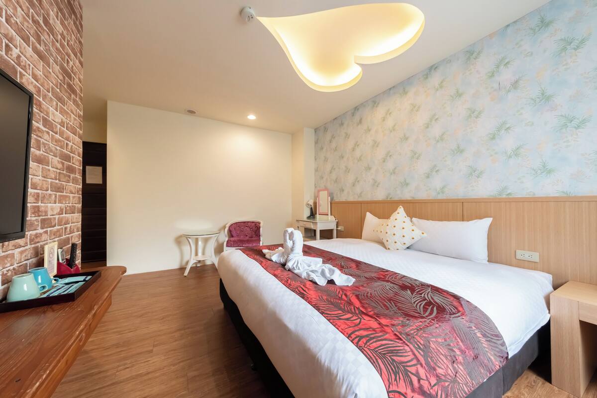Quarto de hotel com uma cama grande, cabeceira de madeira, TV de tela plana, uma mesinha e uma cadeira.