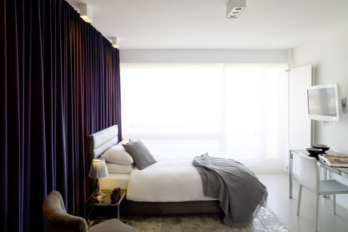 A modern hotel room with a large bed, a bedside table with a lamp, a chair, and a TV mounted on the wall.