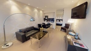 Ein modernes Wohnzimmer mit Sofa, Couchtisch und Stehlampe. Es gibt einen Essbereich mit Stühlen und einem Tisch, und im Hintergrund eine Küchenzeile.