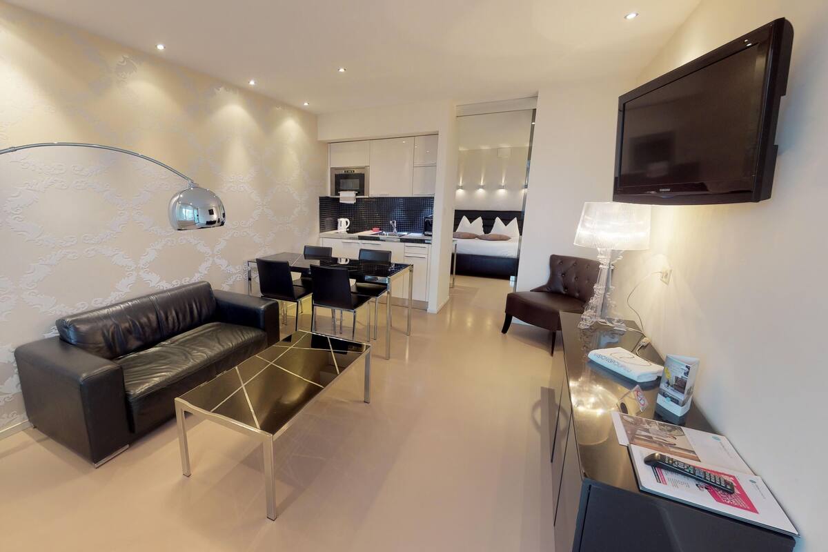A modern living room with a sofa, coffee table, and floor lamp. There is a dining area with chairs and a table, and a kitchenette in the background.