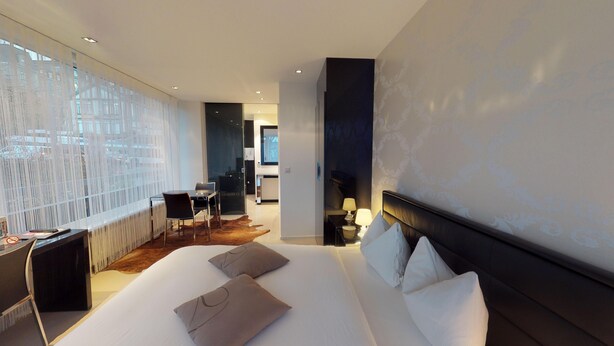 Ein modernes Hotelzimmer mit einem großen Bett, einem Schreibtisch und einem Sessel. Große Fenster bieten einen Blick auf die Stadt.