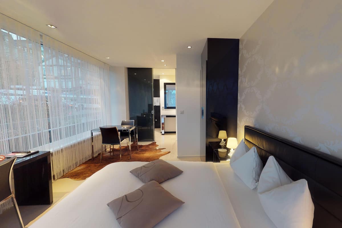A modern hotel room with a large bed, a desk, and a chair. There is a view of the city through large windows.
