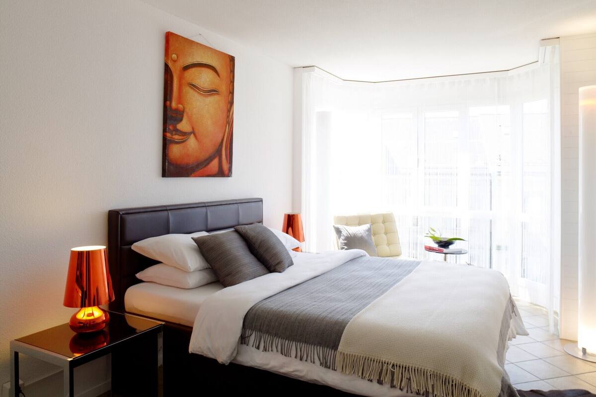 A modern hotel room with a large bed, a bedside table with a lamp, a window with curtains, and a Buddha painting on the wall.