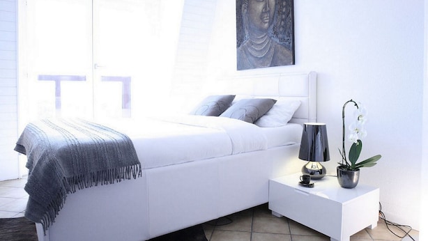 Un dormitorio moderno con una cama blanca, almohadas grises, un plaid gris, una mesita de noche con una lámpara, una pequeña planta en maceta y una pintura de Buda en la pared.