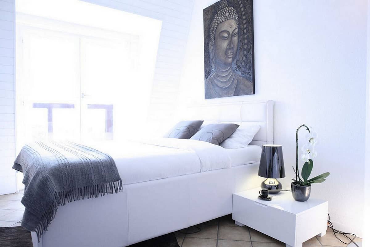 A modern bedroom with a white bed, grey pillows, a grey throw blanket, a bedside table with a lamp, a small potted plant, and a Buddha painting on the wall.