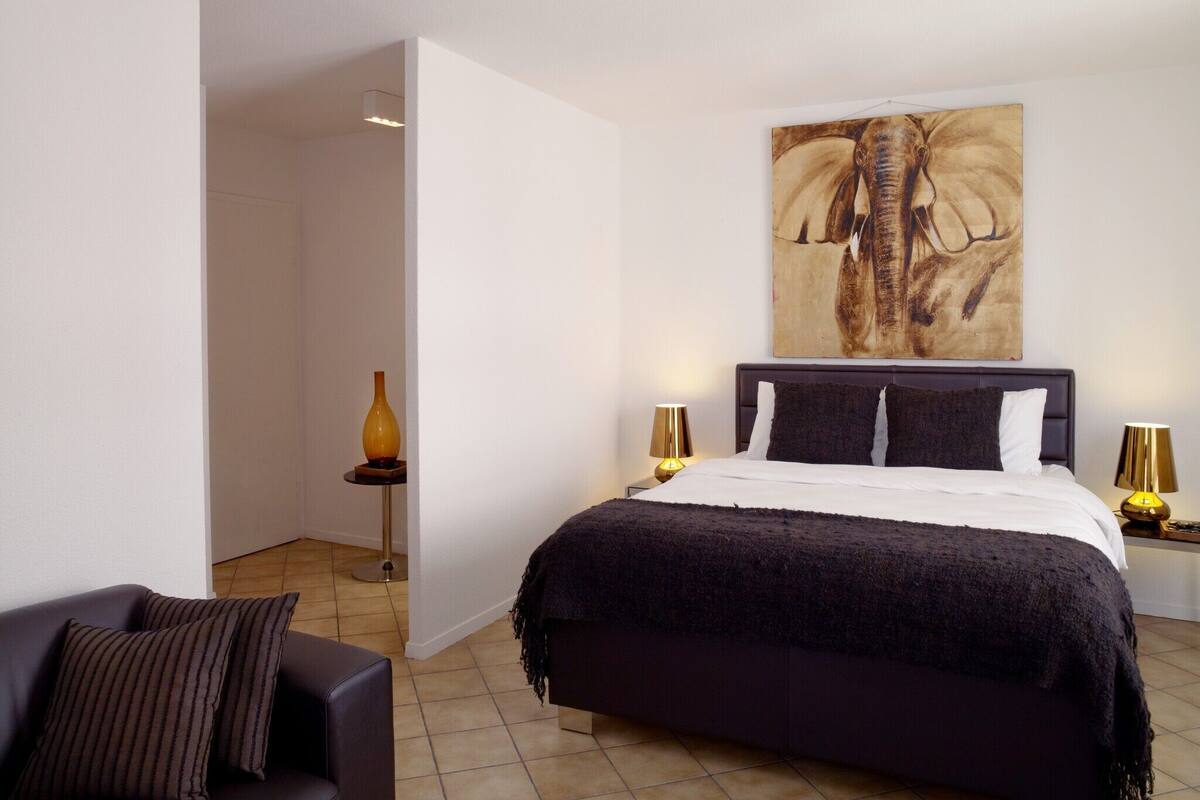 A modern bedroom with a large bed, a dark-colored sofa, a vase, and a wall-mounted elephant painting.