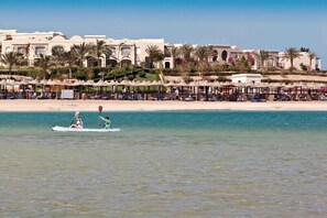 A beach resort with a row of beach umbrellas and lounge chairs along the shore, a couple kayaking, and a building with arches and palm trees in the background. - TUI MAGIC LIFE KALAWY - All inclusive (El Quseir)