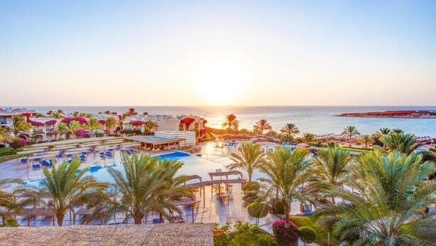 Een resort met een groot zwembad, palmbomen en uitzicht op de oceaan bij zonsondergang.
