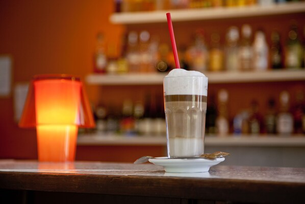 A clear glass with a straw, a spoon, and a cookie on a white plate, with a blurred background of shelves and bottles. - Holiday Hotel Csopak (Csopak)