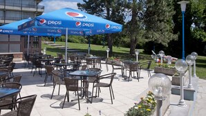 An outdoor seating area with tables and chairs, a large blue Pepsi umbrella, and a building in the background. - Holiday Hotel Csopak (Csopak)