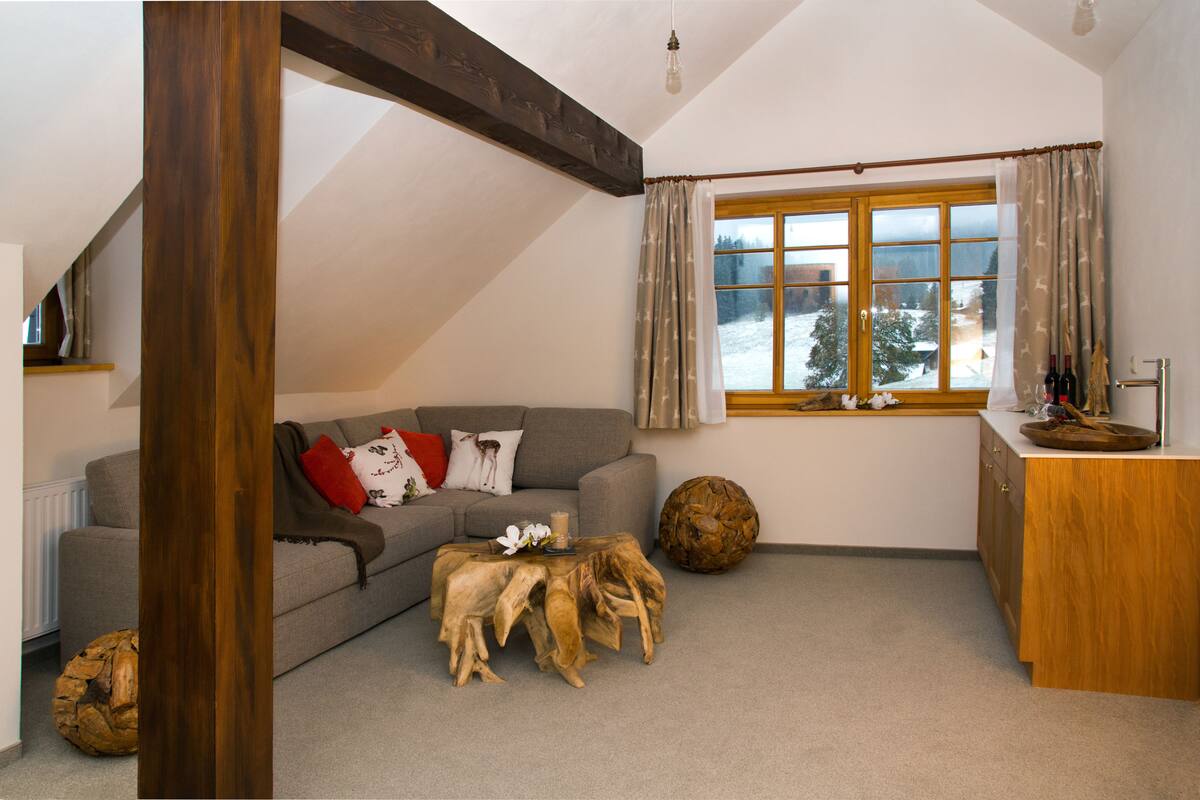 Ein gemütliches Wohnzimmer mit grauem Sofa, Couchtisch aus Holz und einem großen Fenster mit Vorhängen.