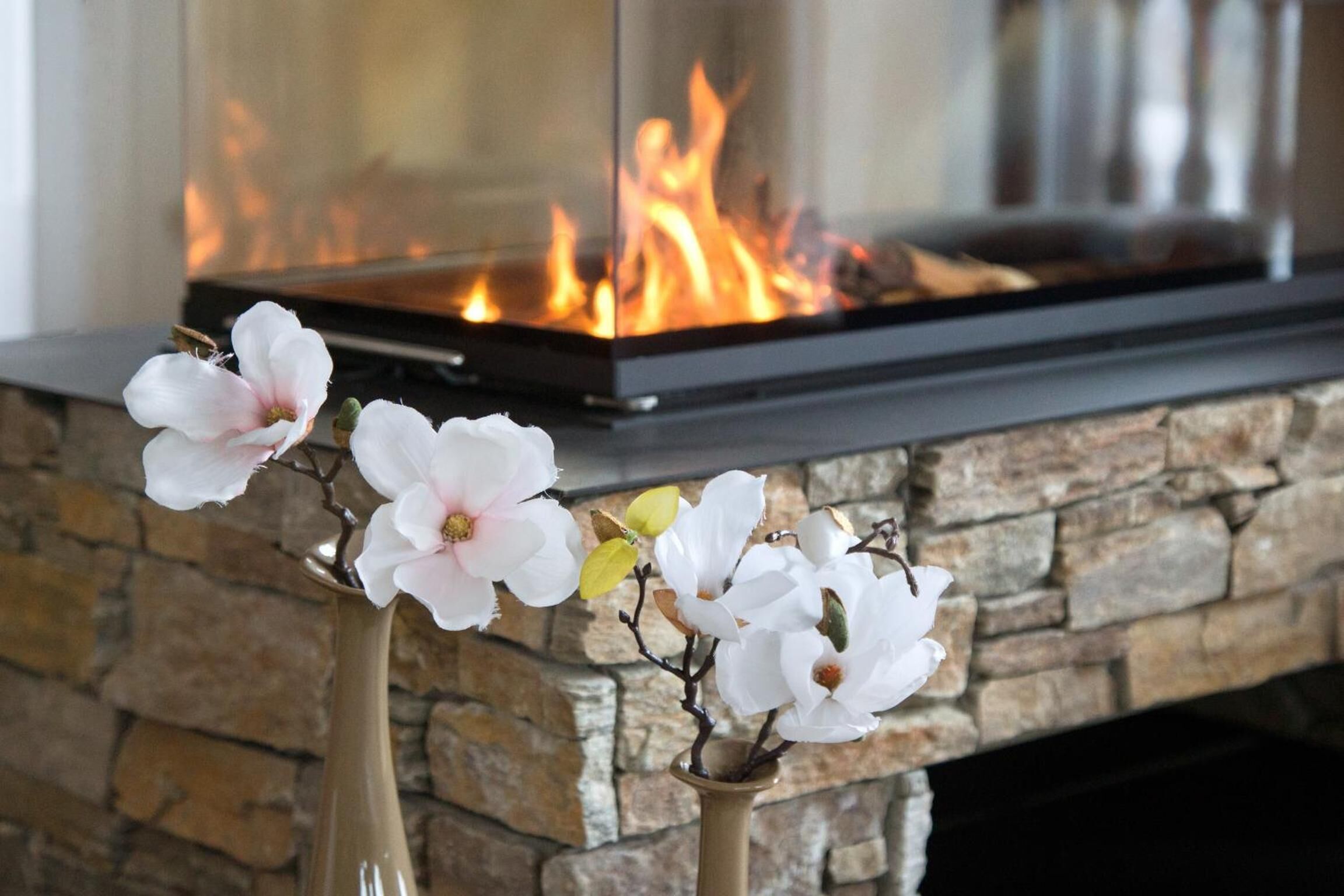 Une cheminée en pierre avec une façade en verre moderne, et trois fleurs blanches dans un vase posé sur le manteau de la cheminée.