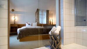 A modern bedroom with a glass partition, a wooden bed frame, a bedside table with a lamp, and a decorative wooden structure.