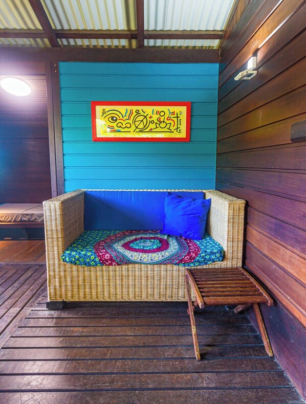 Un sofá de mimbre con un cojín colorido, un banco de madera y una pared con una obra de arte enmarcada.