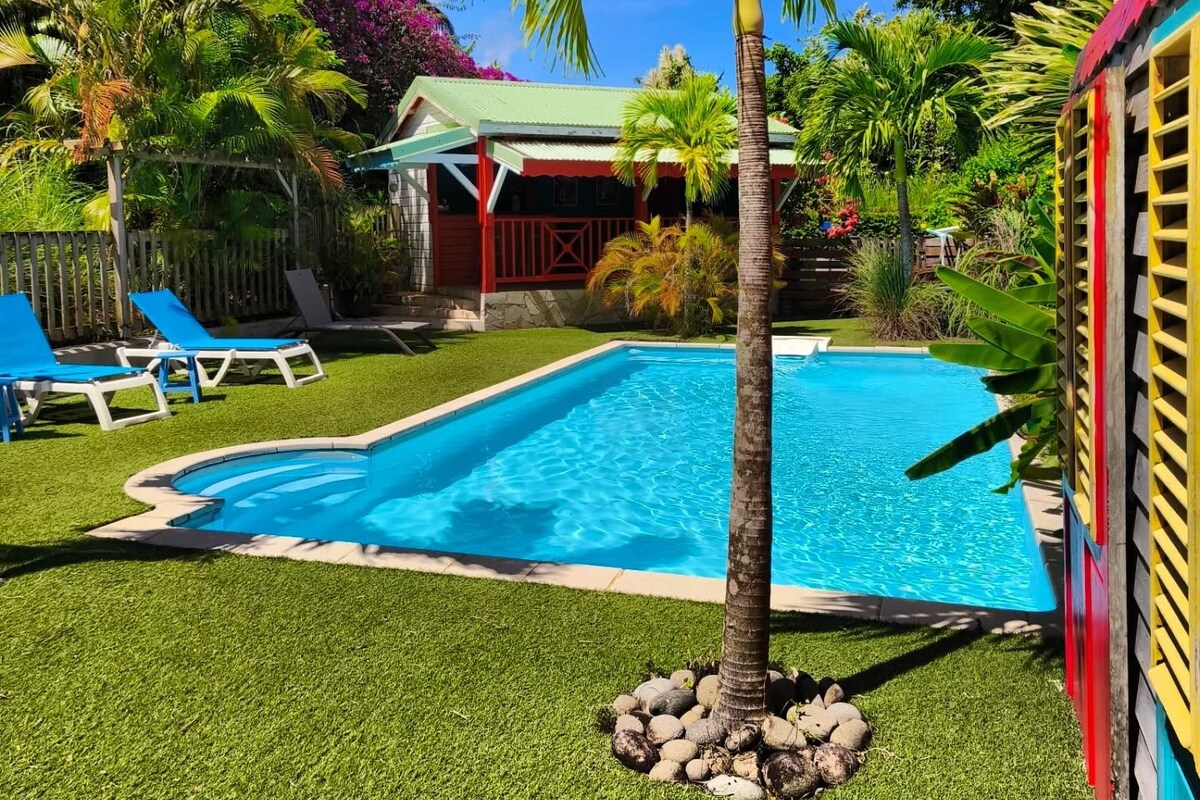 A swimming pool surrounded by artificial grass, palm trees, and a small building with a red roof.