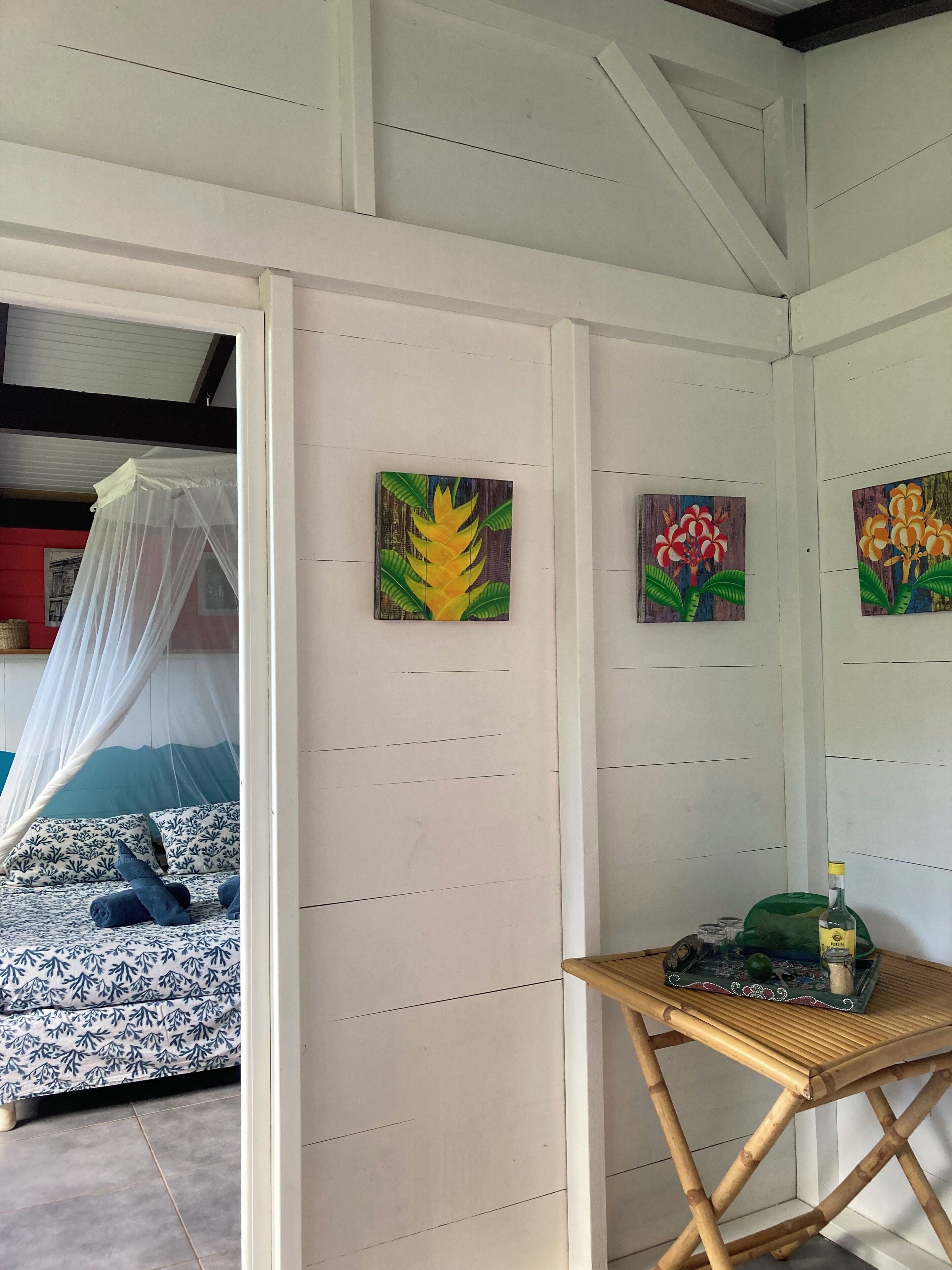 A room with a bed, a table with a bottle, and three paintings on the wall.