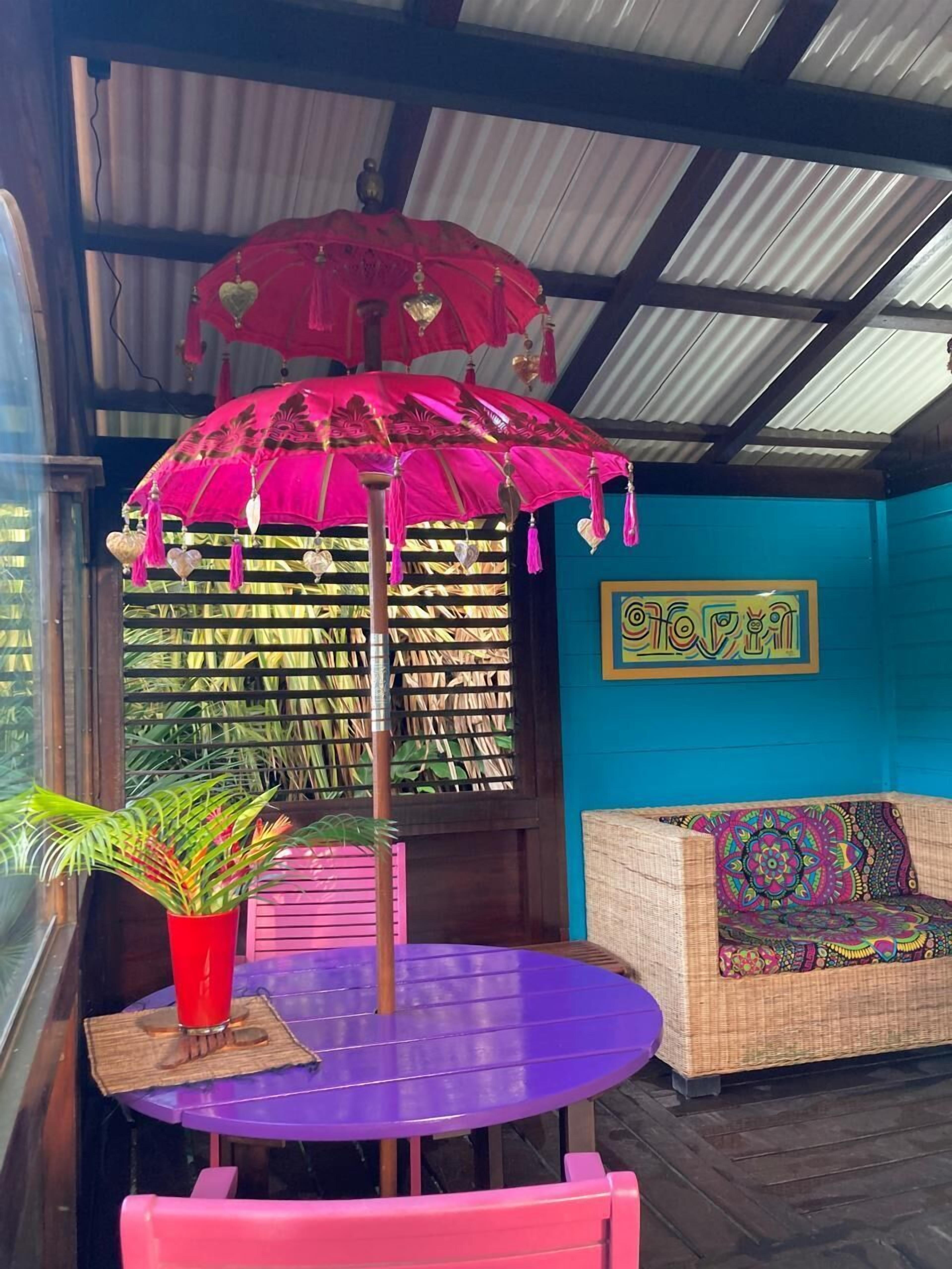 A room with a purple table, a red umbrella, a wicker sofa with colorful cushions, and a wall with a framed artwork.