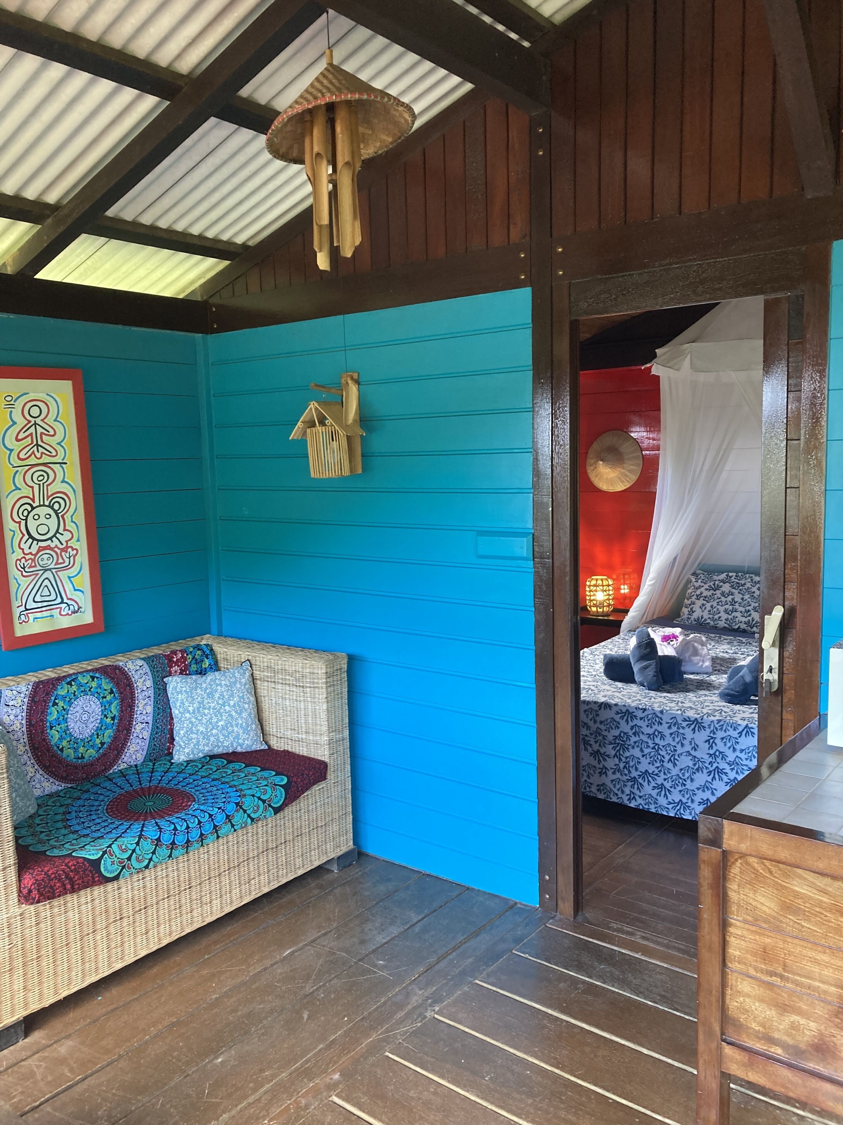 Quarto com parede azul, sofá de vime com almofadas estampadas, cama com dossel e luminária pendente.