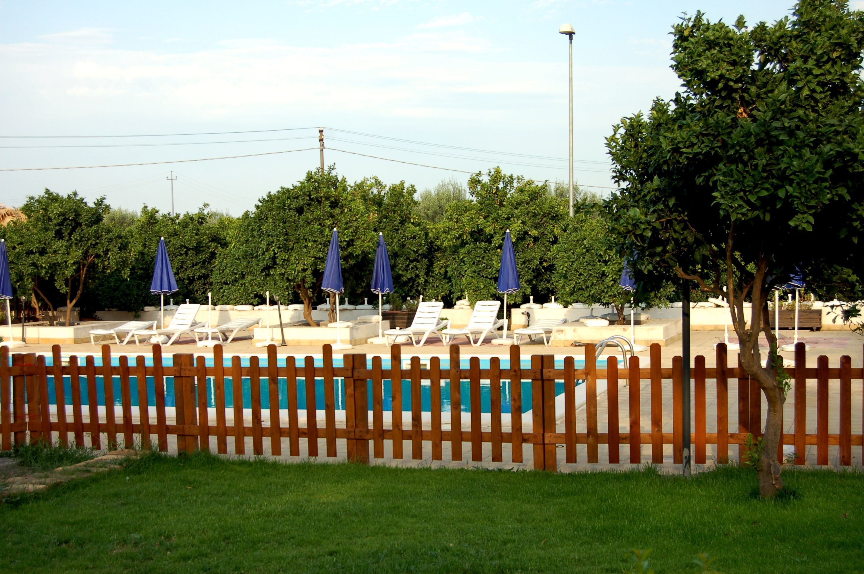 Area piscina con lettini e ombrelloni, circondata da una recinzione in legno e alberi.