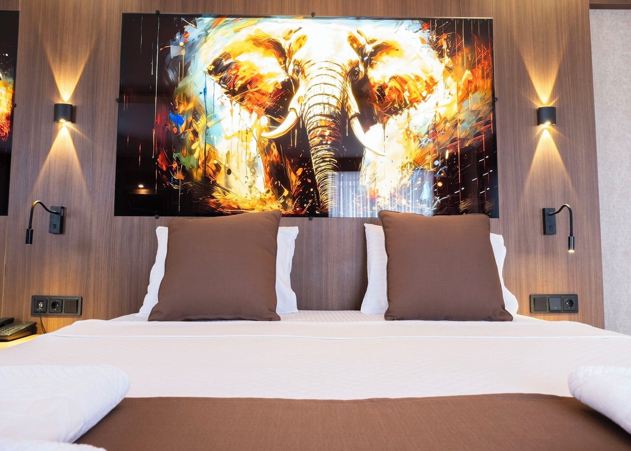 Quarto de hotel com uma cama grande, duas almofadas castanhas, uma TV de tela plana com uma pintura de elefante e luminárias fixadas na parede.