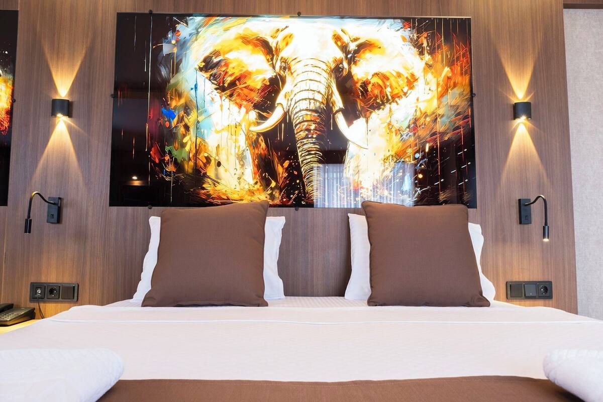 Quarto de hotel com uma cama grande, duas almofadas castanhas, uma TV de tela plana com uma pintura de elefante e luminárias fixadas na parede.
