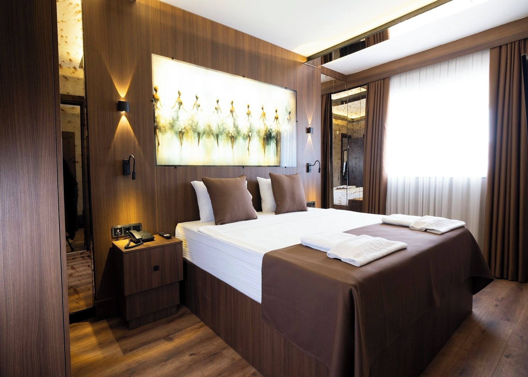 Quarto de hotel moderno com cama grande, mesas de cabeceira, uma obra de arte na parede e uma janela ampla com cortinas.