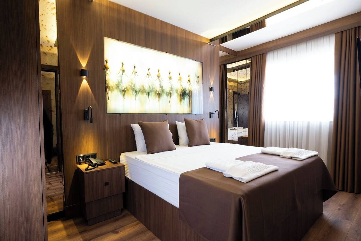 Quarto de hotel moderno com cama grande, mesas de cabeceira, uma obra de arte na parede e uma janela ampla com cortinas.