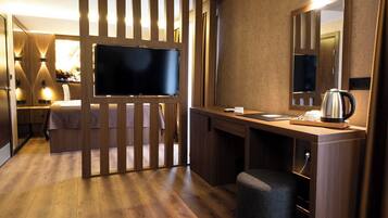 A hotel room with a wooden floor, a bed with a white headboard, a desk with a kettle, a flat-screen TV, and a mirror.