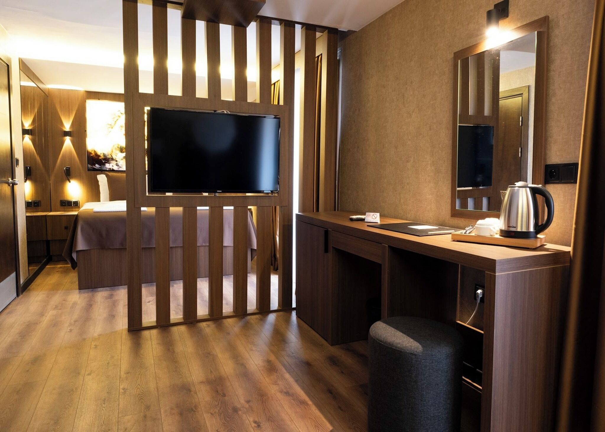 Quarto de hotel com secretária de madeira, televisão de tela plana, espelho e área de cozinha compacta.