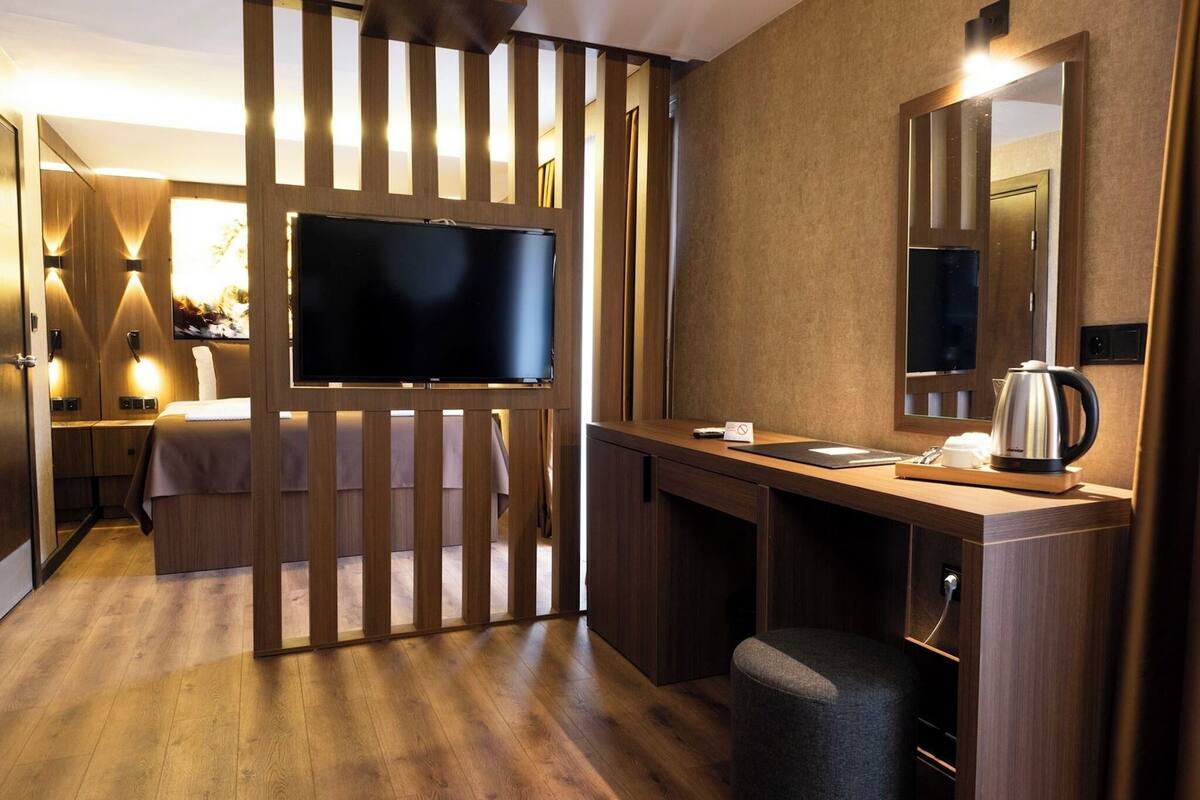 Quarto de hotel com secretária de madeira, televisão de tela plana, espelho e área de cozinha compacta.