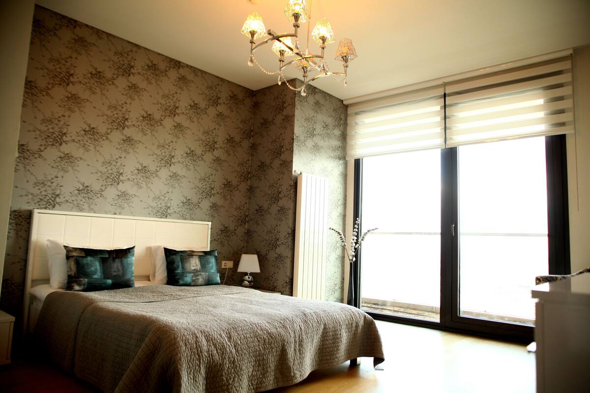 A bedroom with a large bed, a chandelier, a window with blinds, and a nightstand with a lamp.