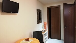 A hotel room with a television mounted on the wall, a dining table with a chair, a wooden door, and a small cabinet with decorative items. - B&B Li Figuli (Lecce)