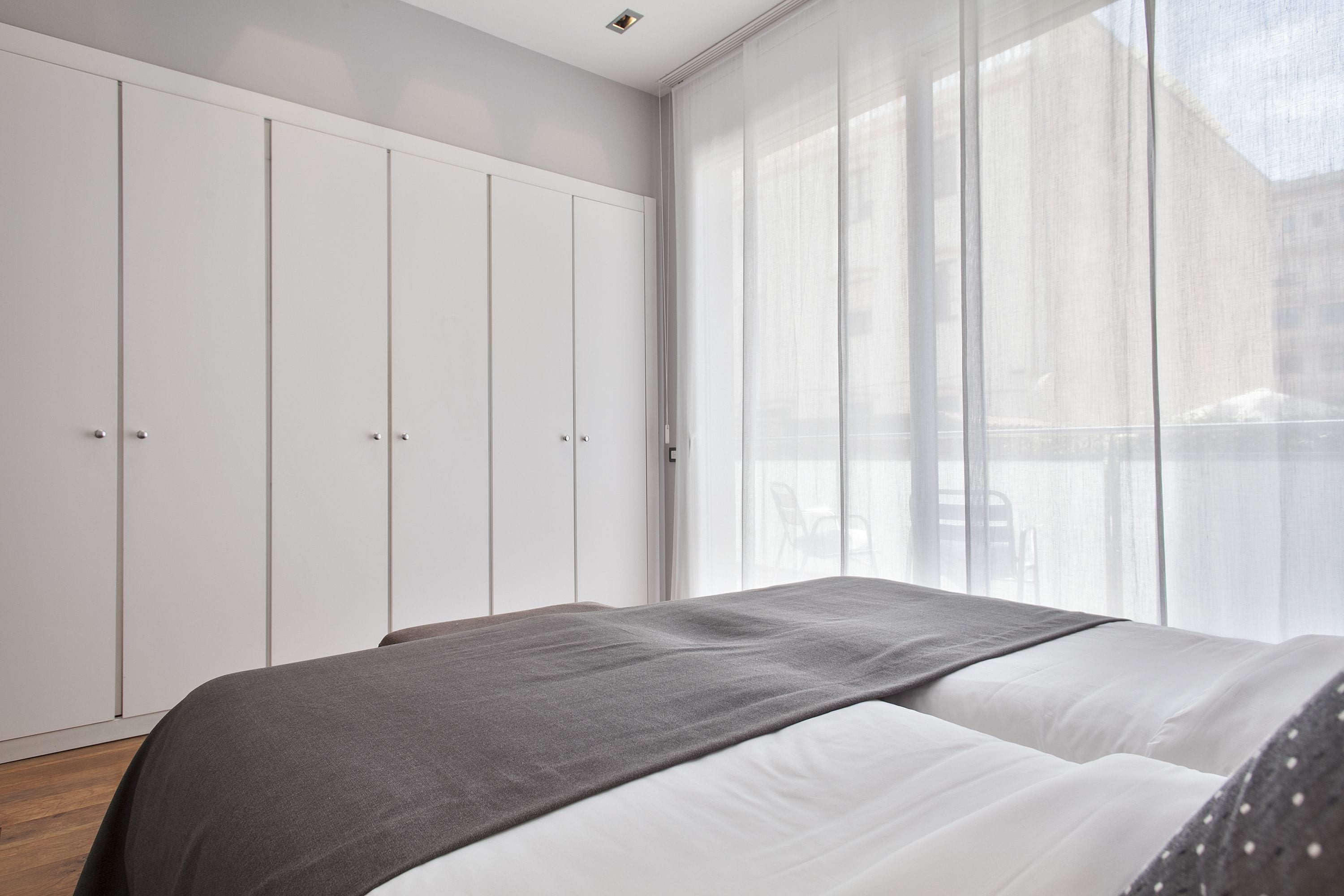 Un dormitorio con cama, armario y un amplio ventanal con cortinas de gasa.