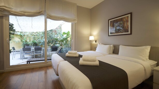 A hotel room with a bed, a bedside table, a lamp, and a view of an outdoor area with plants and a patio. - Bonavista Apartments - Passeig de Gracia (Barcelona)
