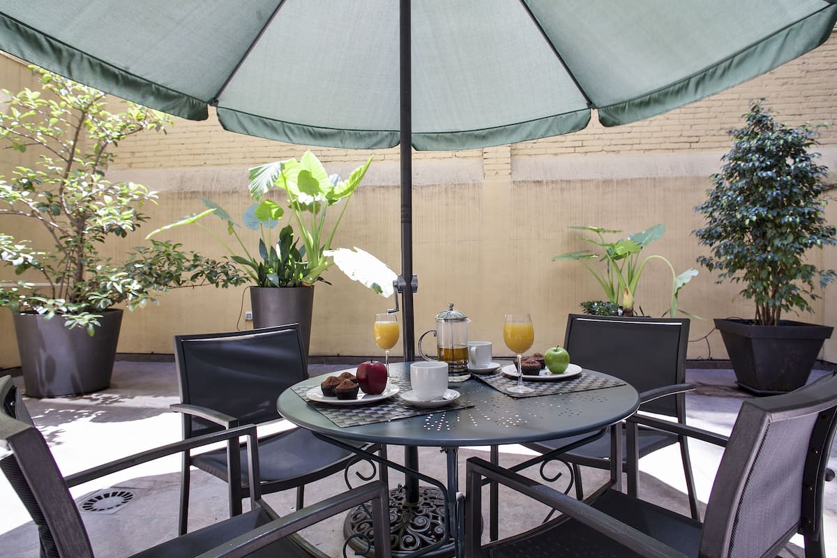 A round table with chairs, a green umbrella, and a breakfast setting including muffins, apples, and orange juice.