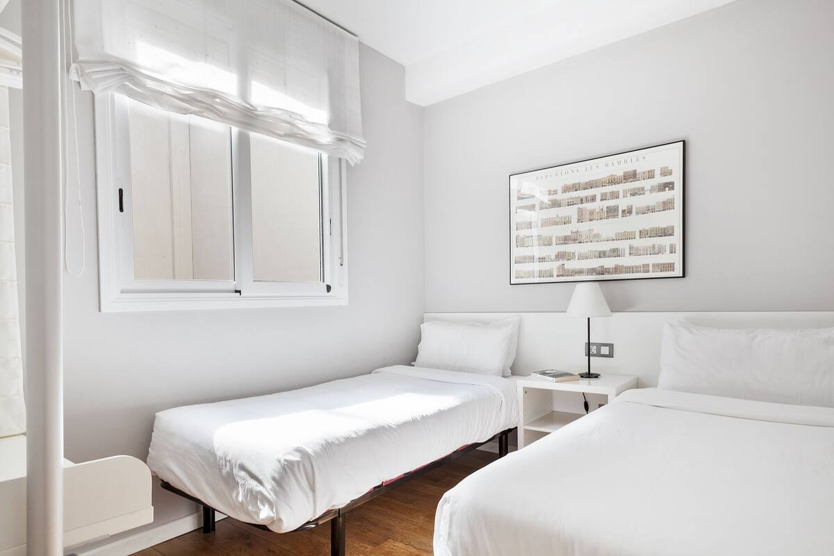 A minimalist hotel room with two single beds, a small bedside table with a lamp, and a large window with sheer curtains.