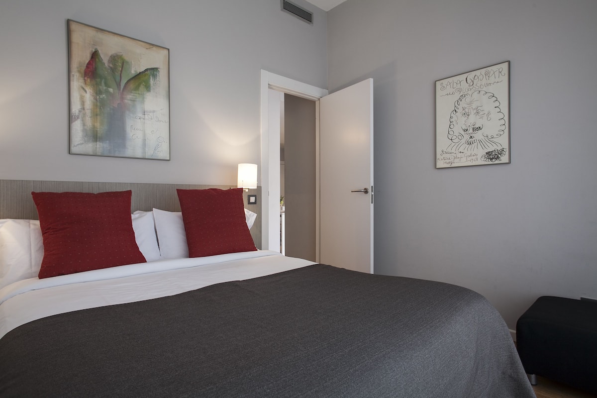 A hotel room with a large bed, red and white pillows, a grey blanket, a wall-mounted lamp, and two framed artworks.