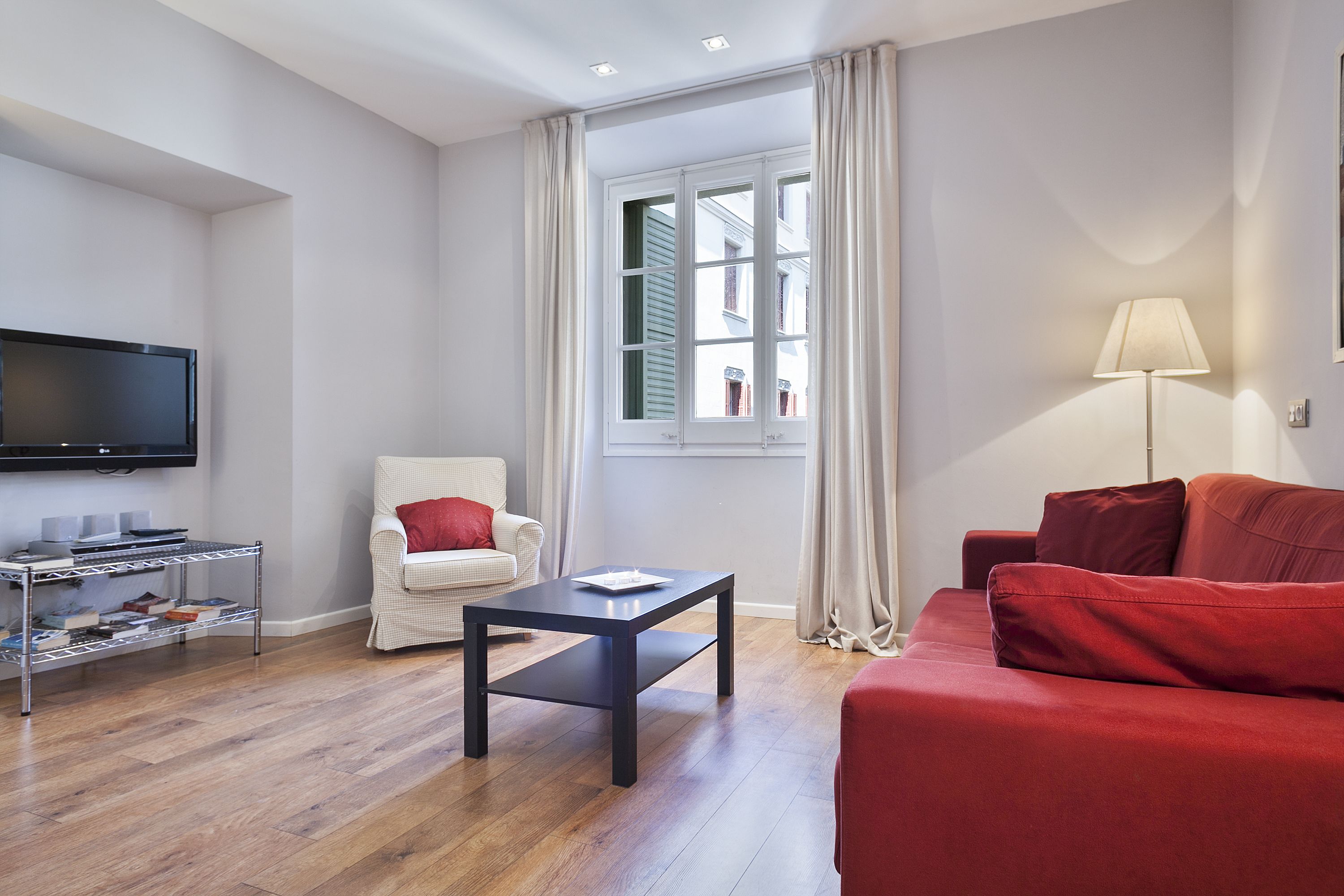A living room with a flat-screen TV, a white armchair, a red sofa, a coffee table, and a window with curtains.