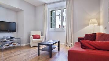 A living room with a flat-screen TV, a white armchair, a red sofa, a coffee table, and a window with curtains.