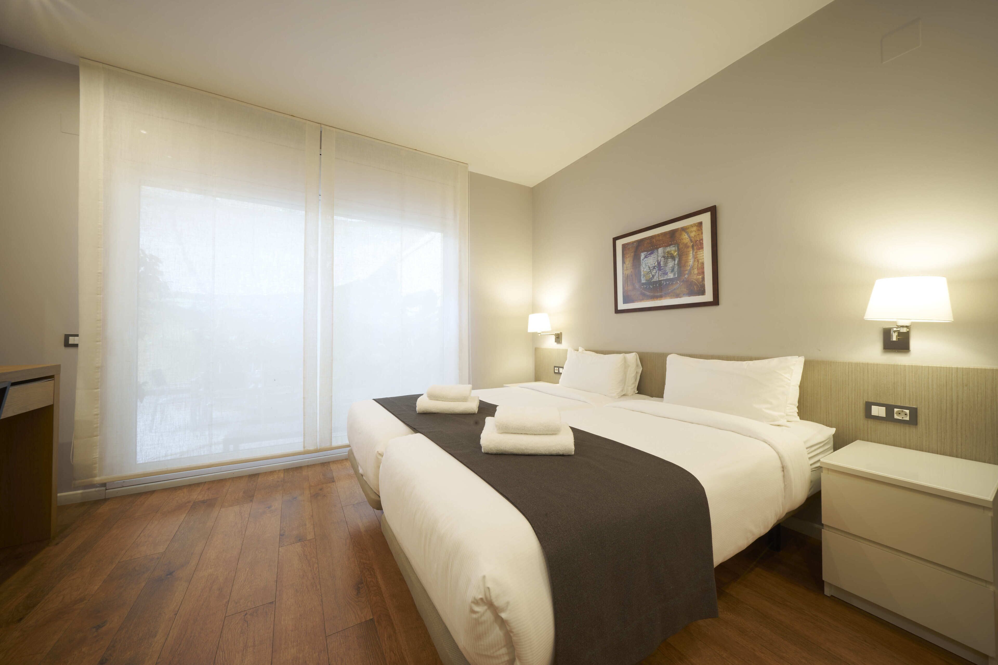 A hotel room with a large bed, a bedside table, a nightstand, a window with curtains, and a wall-mounted picture.