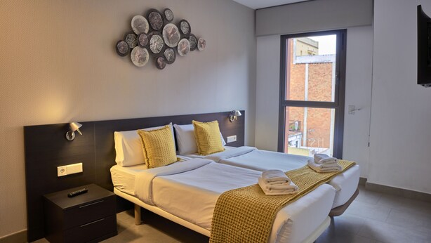 A modern hotel room with two beds, a wall-mounted artwork, a nightstand, and a window with a view of a building. - Bonavista Apartments - Virreina (Barcelona)