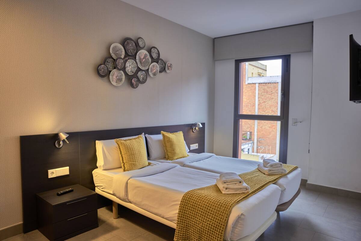 A modern hotel room with two beds, a wall-mounted artwork, a nightstand, and a window with a view of a building.